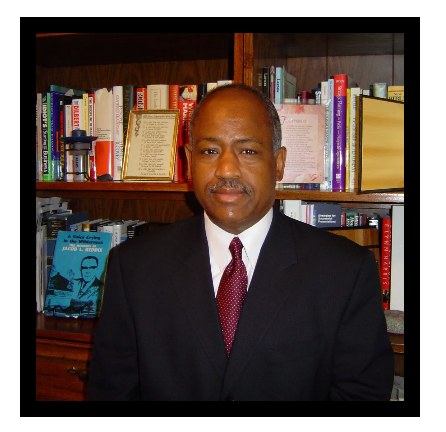 A man in a suit and tie stands in front of a bookshelf filled with books