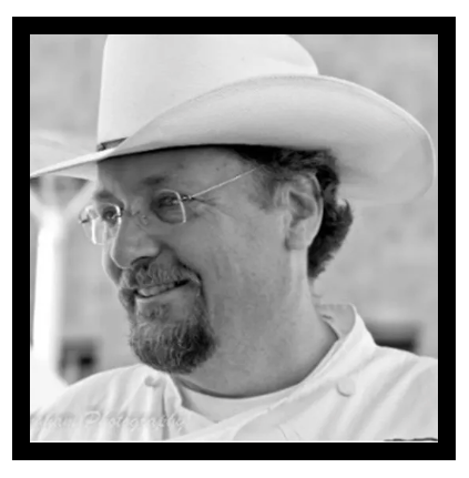 A man wearing a cowboy hat and glasses is smiling in a black and white photo.