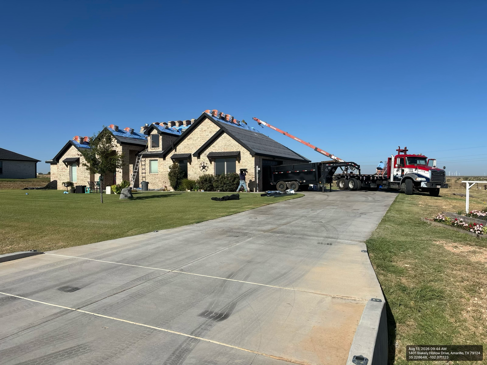 Suburban house under construction with roof trusses, red crane, and a wide concrete driveway under a blue sky