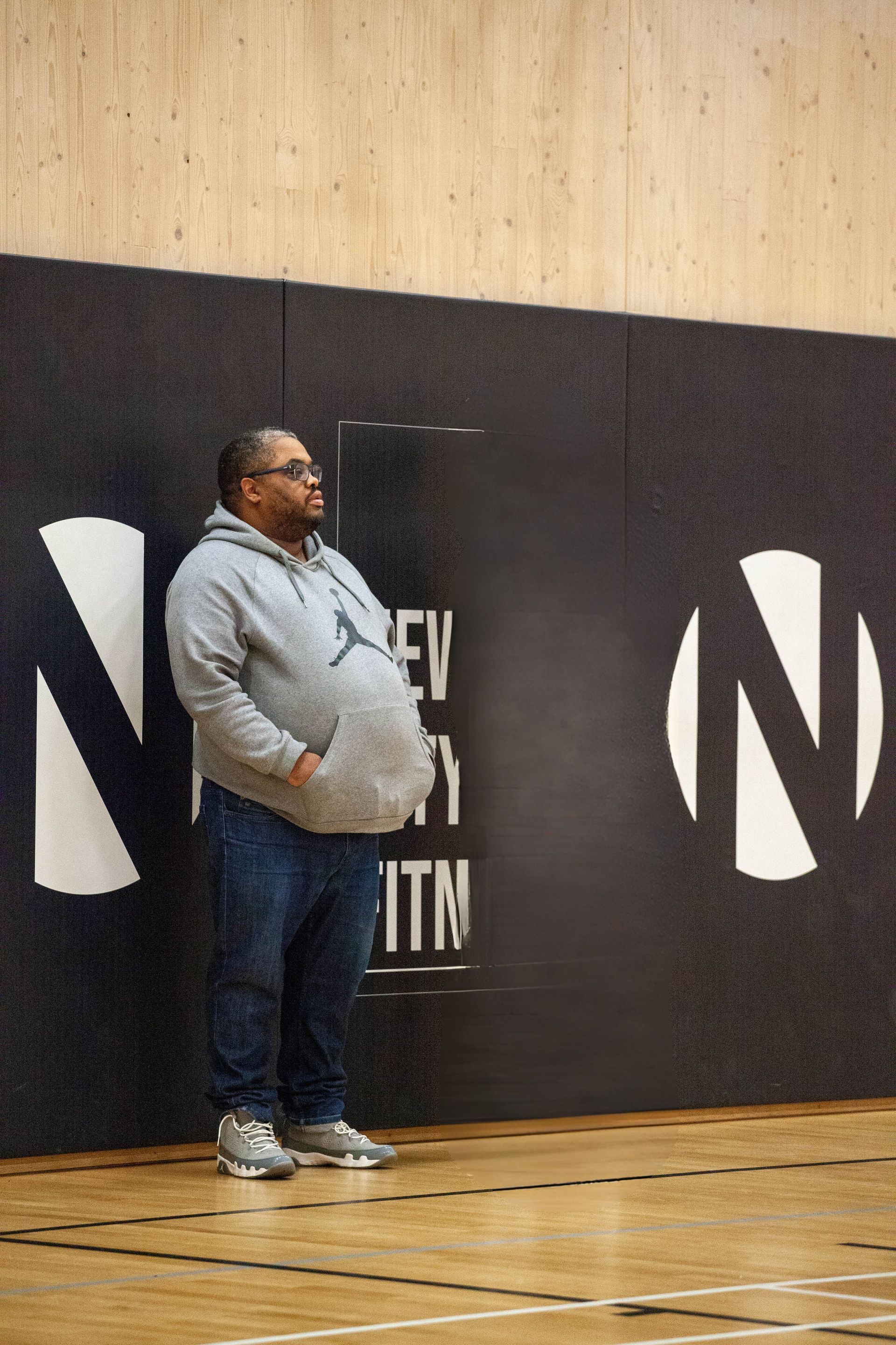 Man in gray hoodie and jeans standing in front of a wall with an 