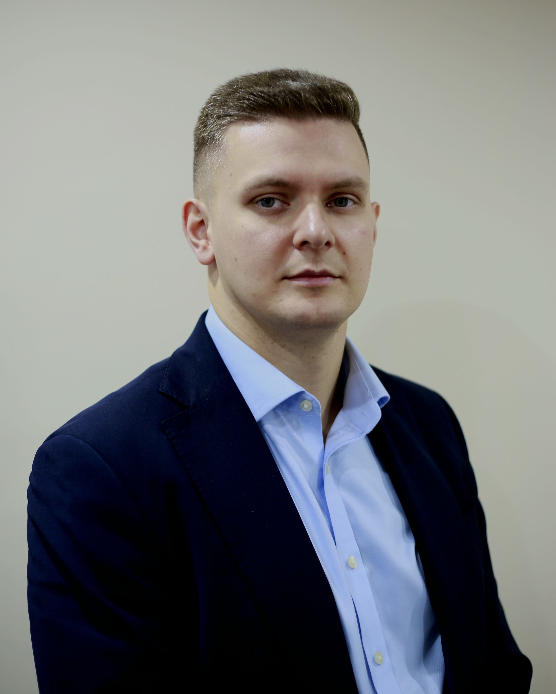 Man in blue shirt and navy blazer looks directly at the camera. Neutral background.