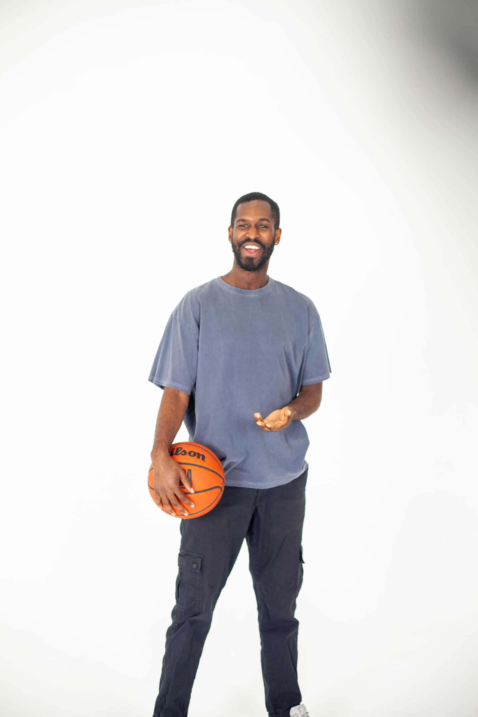 Man smiles, holds basketball, and gestures with one hand against a white backdrop. He wears a blue t-shirt and dark pants.