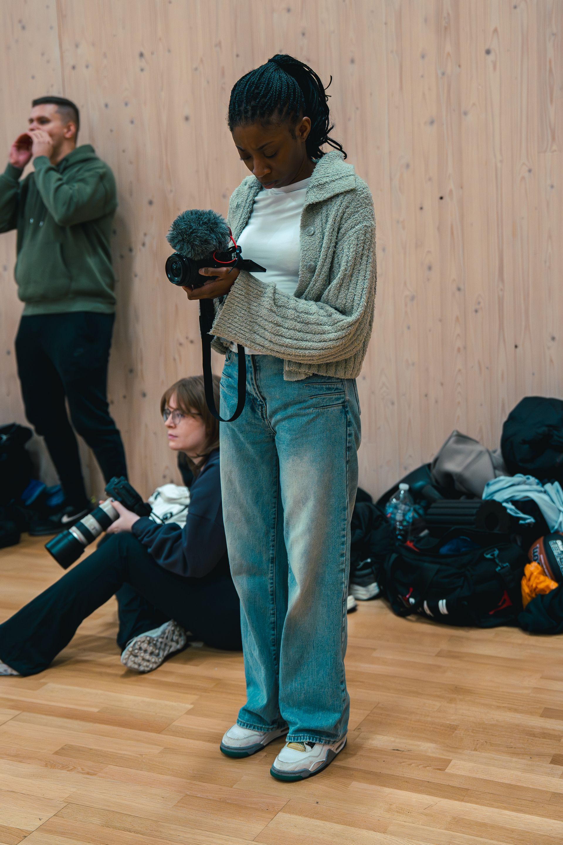 Woman holding a video camera with a microphone, wearing jeans and a sweater. Others are in the background.