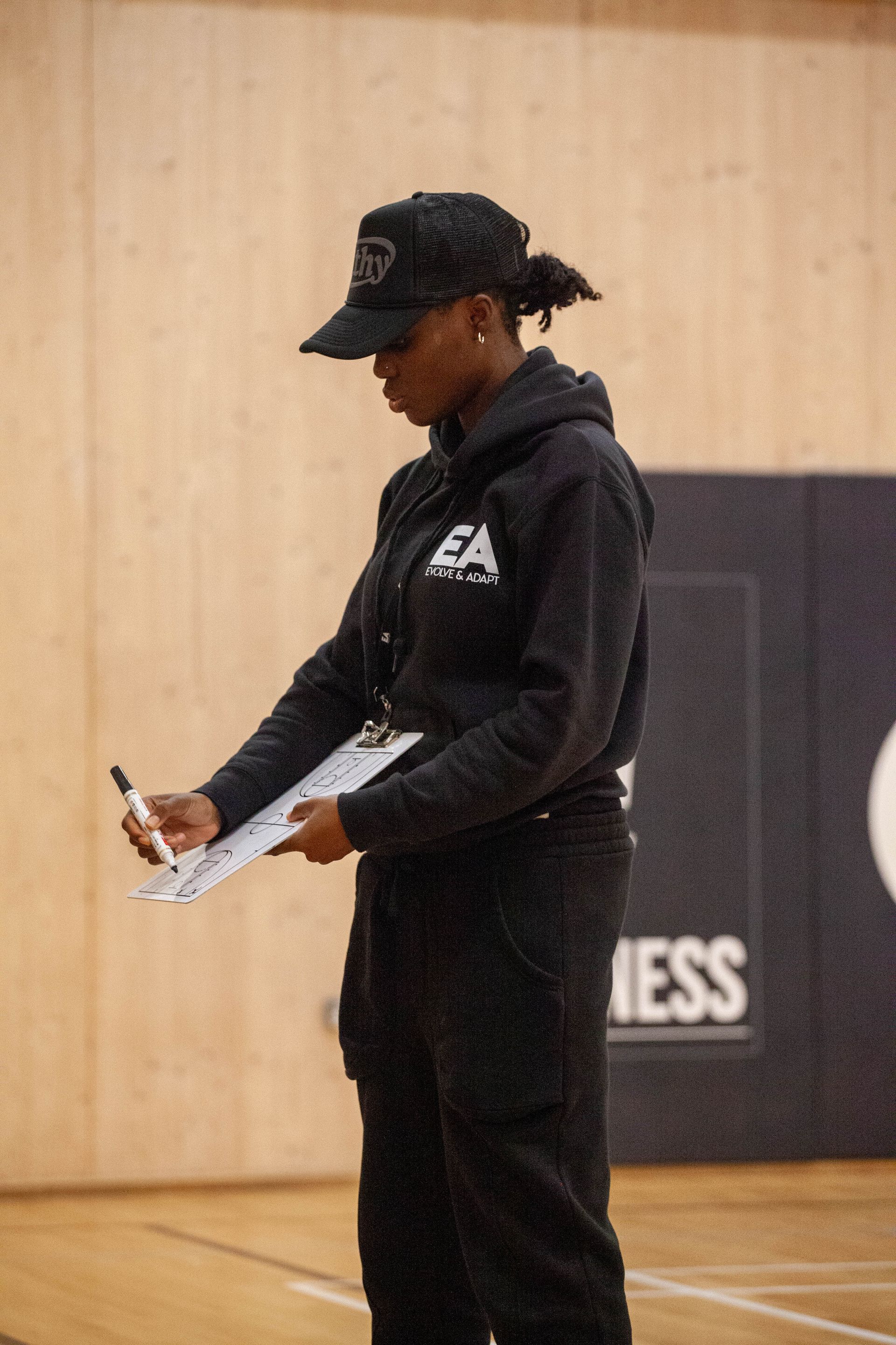Person in black outfit, hat, and hoodie, writing on clipboard in a gym.