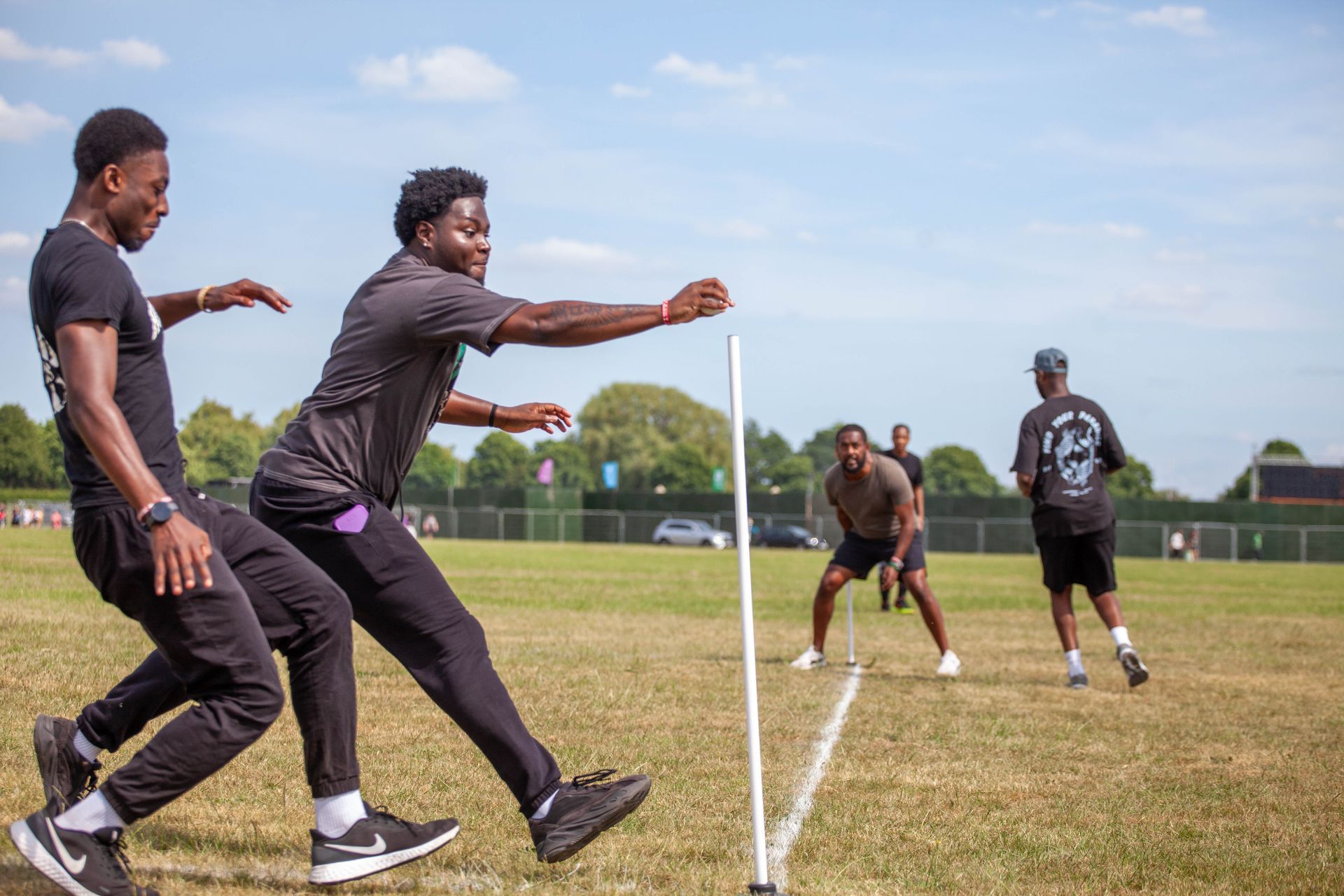 Two young men doing an agility drill on a grassy field, near a white line and other people in the background.