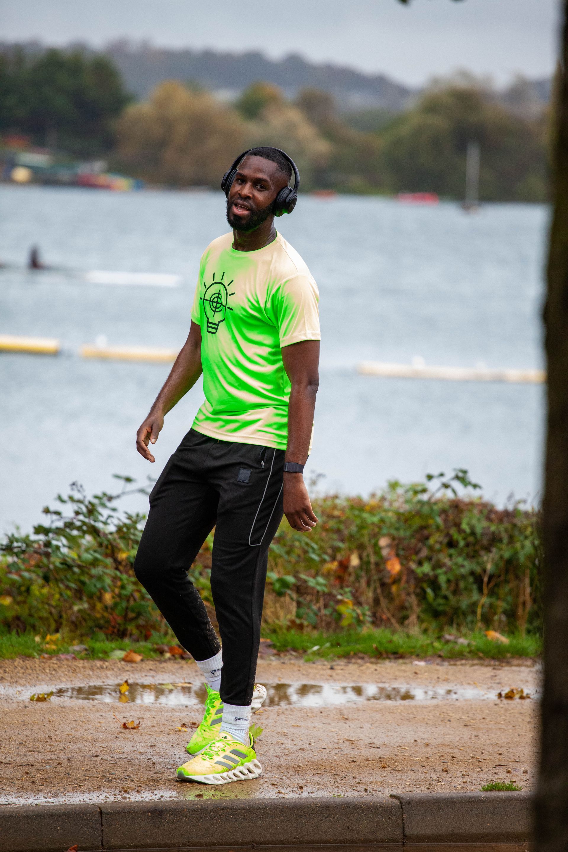 Man in bright green shirt and shoes walks by a lake, wearing headphones, black pants.