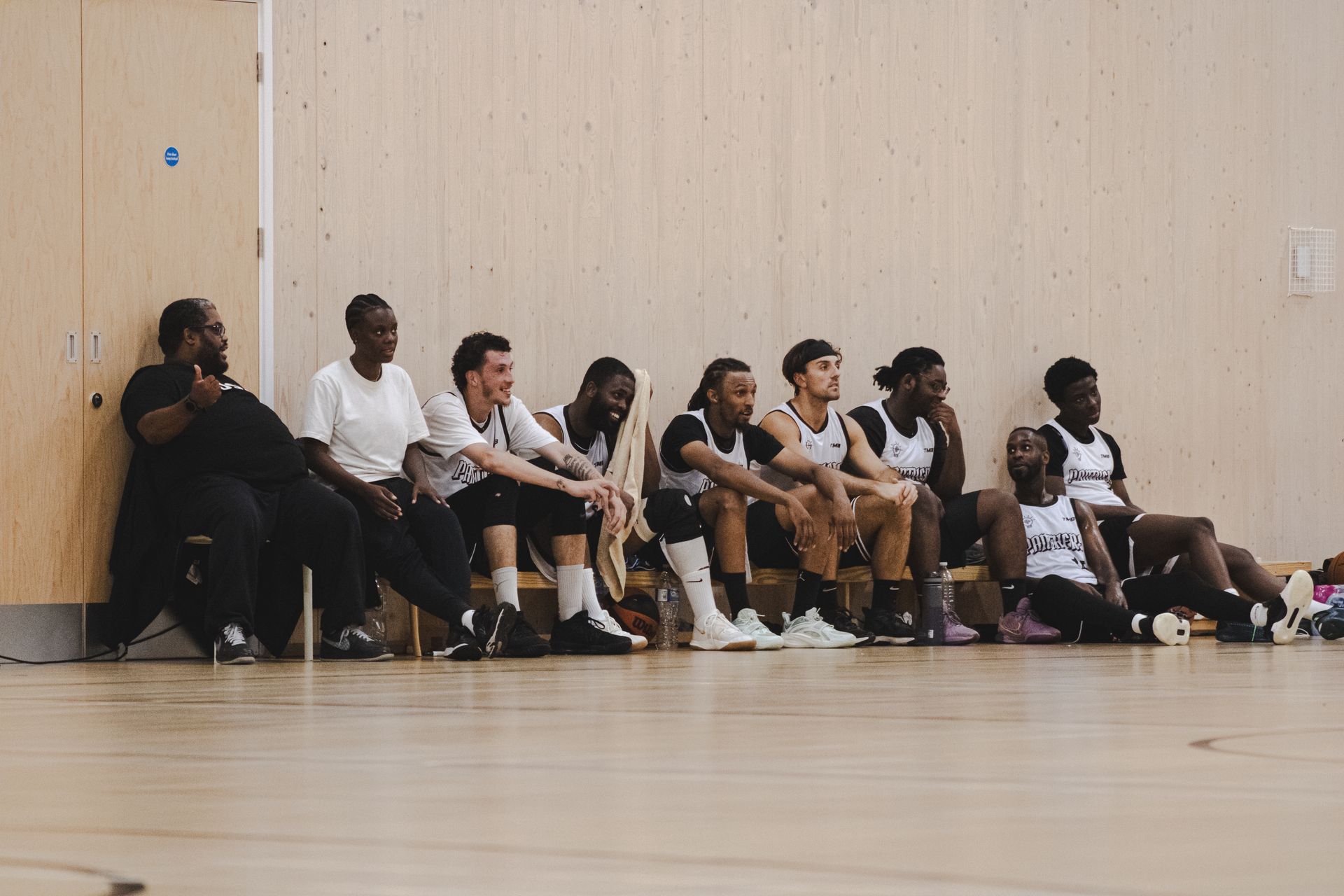 Basketball team on a bench, wooden walls. Players wear white and black jerseys, some looking at the game.