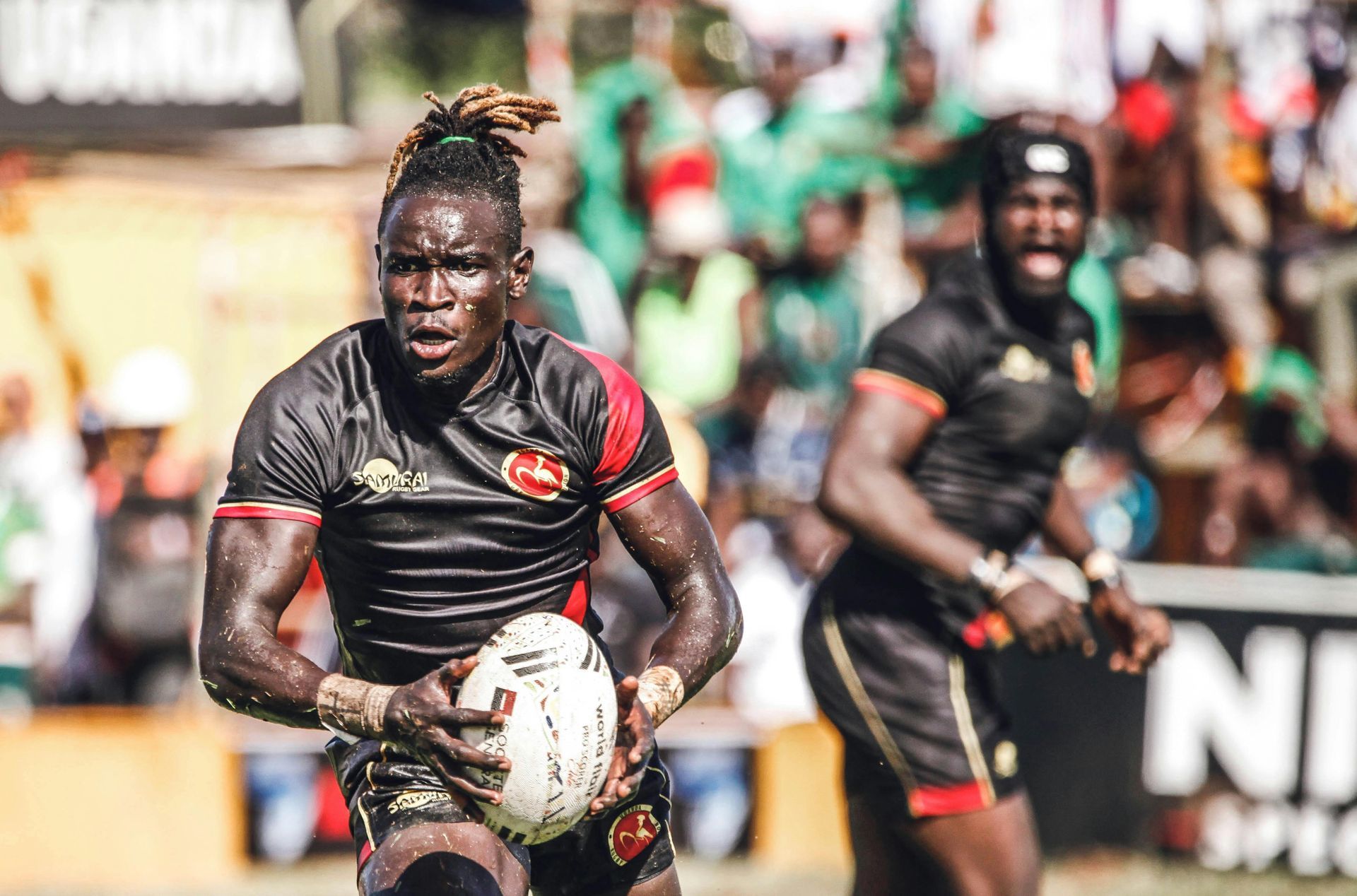 Rugby players in black jerseys with red accents; one running with the ball, other in background cheering.