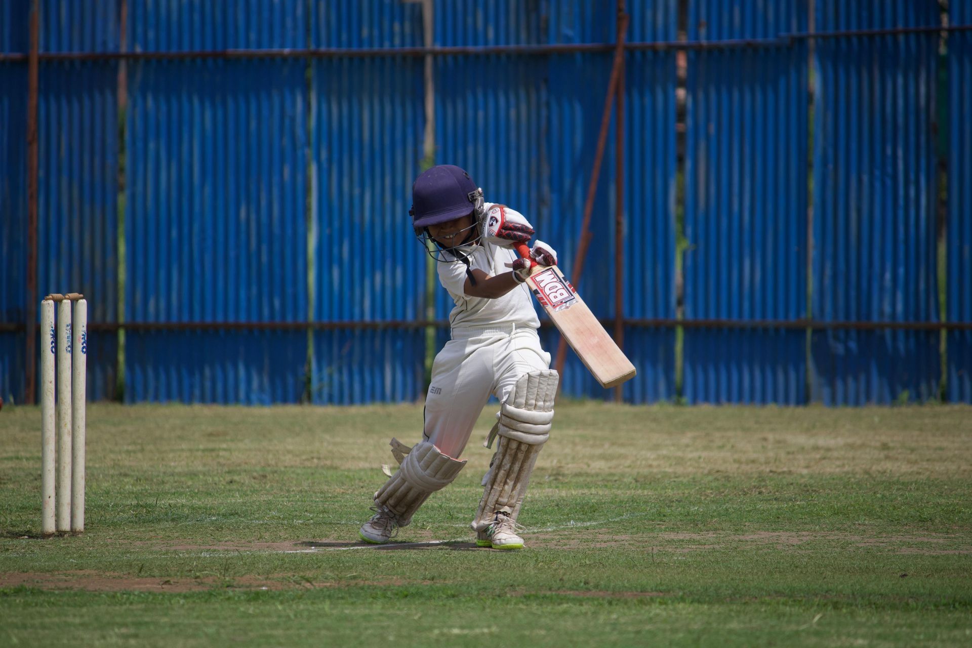 Child batting in a cricket match; wearing white uniform, helmet, swinging bat, on green grass field.