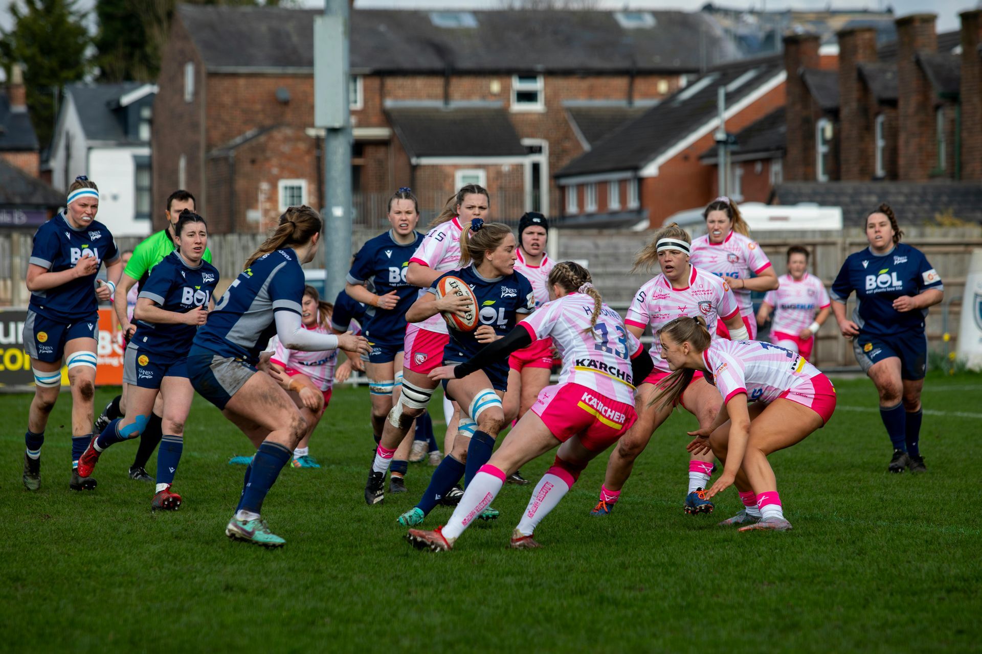 Women's rugby players in blue and pink uniforms competing on a green field. One player runs with the ball, tackled by others.