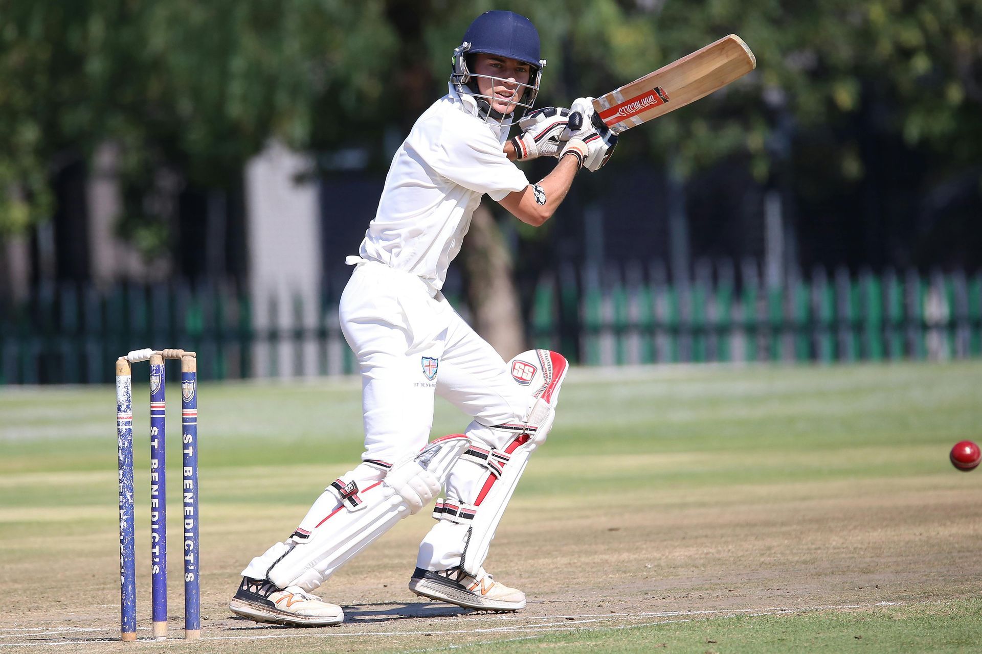 Cricket batter in white uniform swings bat, red ball in flight on sunny field.
