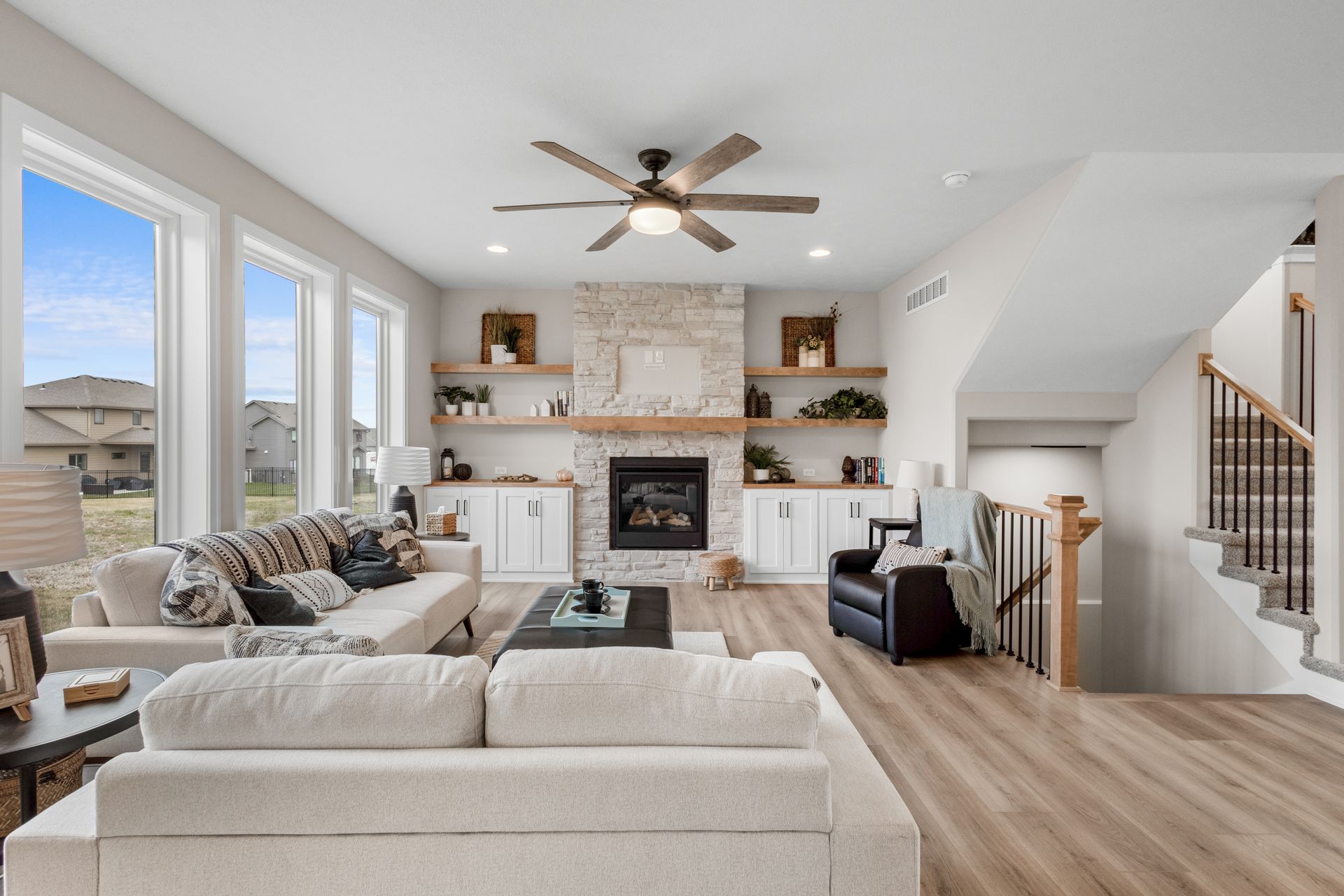 Bright living room with beige sectional, fireplace, floating shelves, and staircase beside large windows