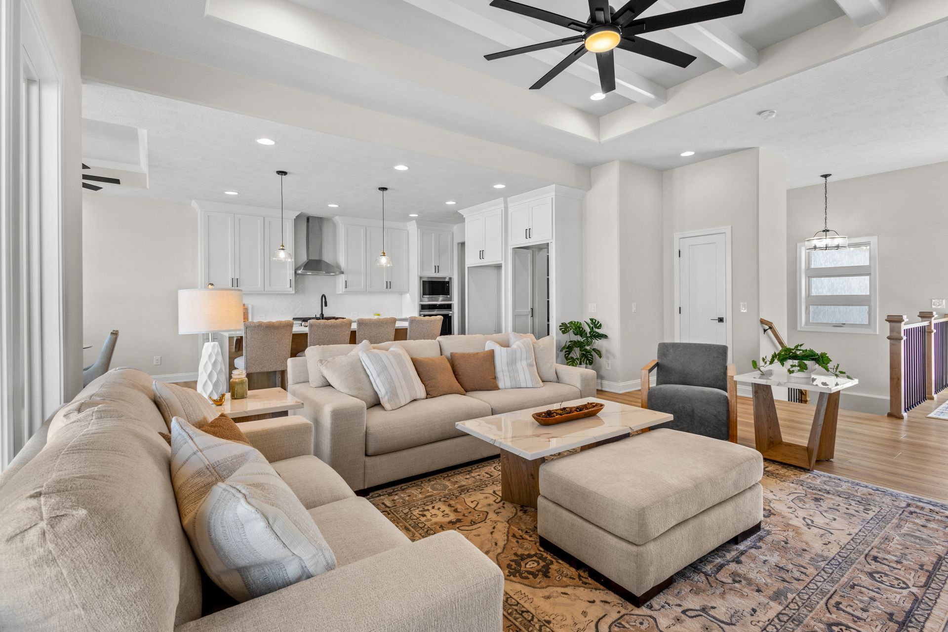 Bright modern living room with beige sectional, ottoman, patterned rug, and open kitchen in the background