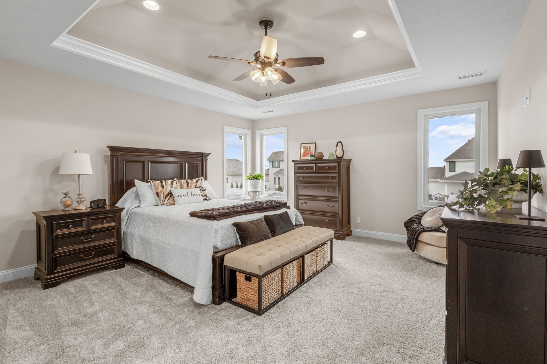 Spacious bedroom with a dark wood bed, gray carpet, bench, dressers, and tray ceiling with a ceiling fan