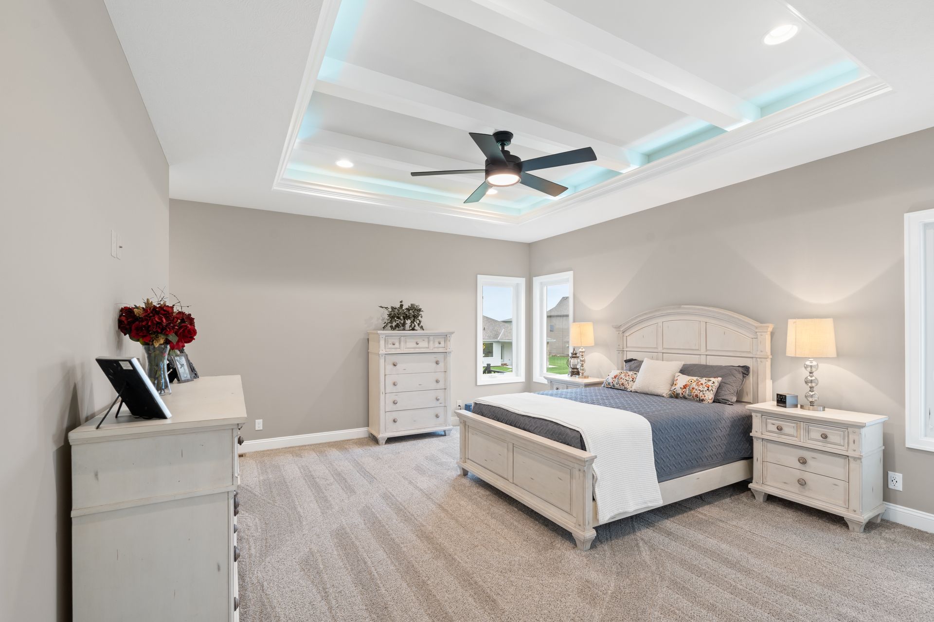 Bright modern bedroom with white furniture, gray carpet, and a tray ceiling with a ceiling fan.
