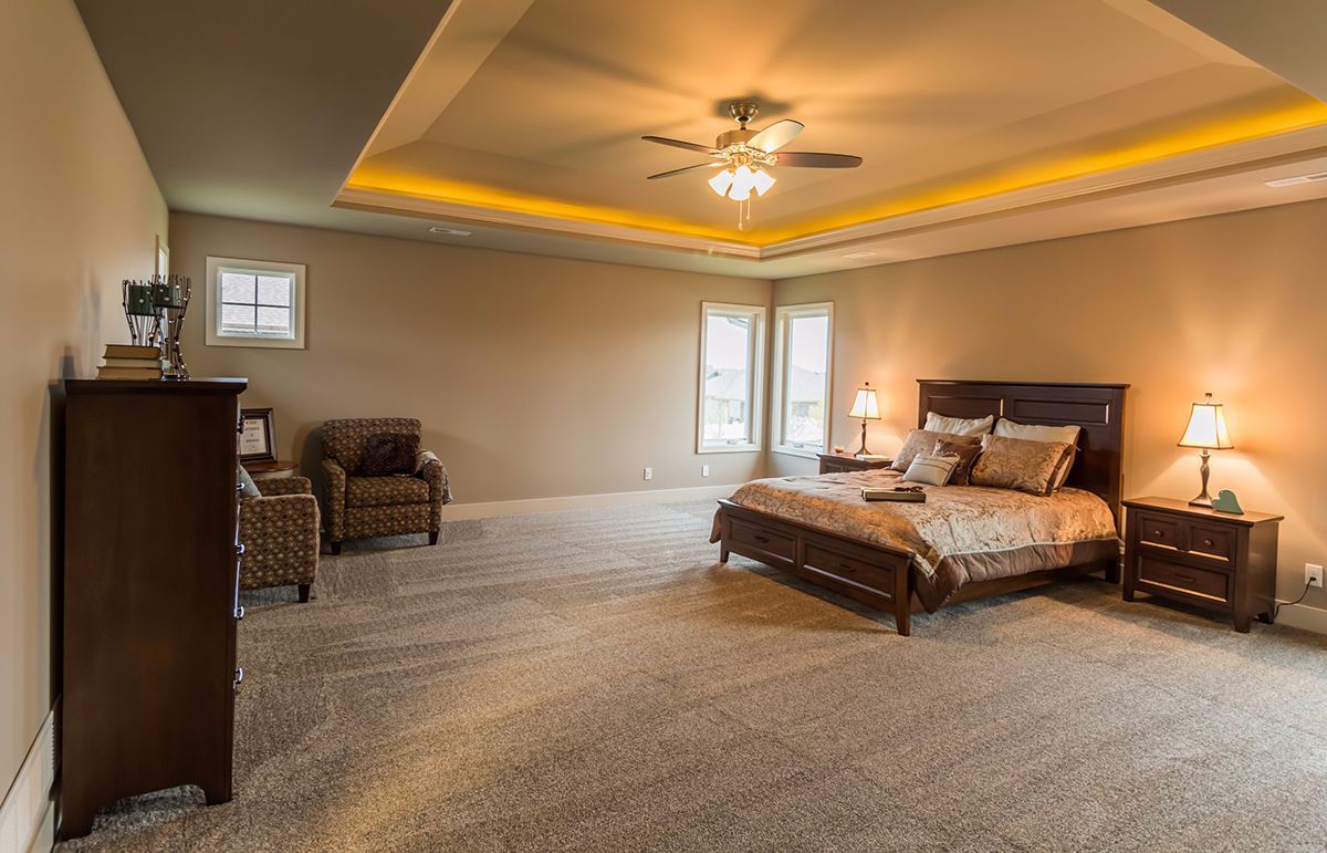 Spacious beige bedroom with a large bed, armchair, dresser, and warm recessed ceiling lighting.