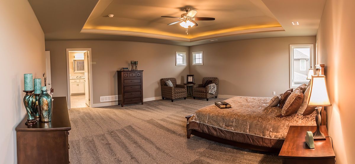 Spacious beige bedroom with tray ceiling, king bed, dresser, loveseat, and nightstand lamp