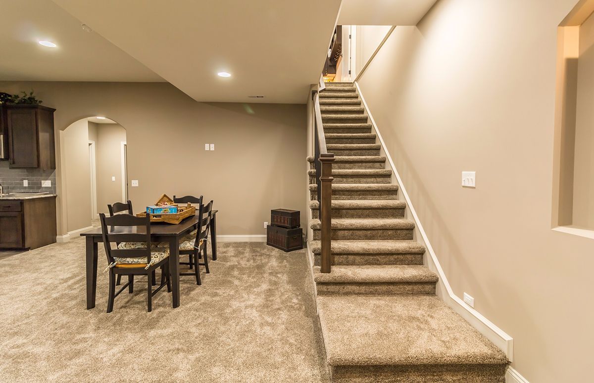 Finished basement with carpeted stairs, dining table, and kitchenette area in beige walls.