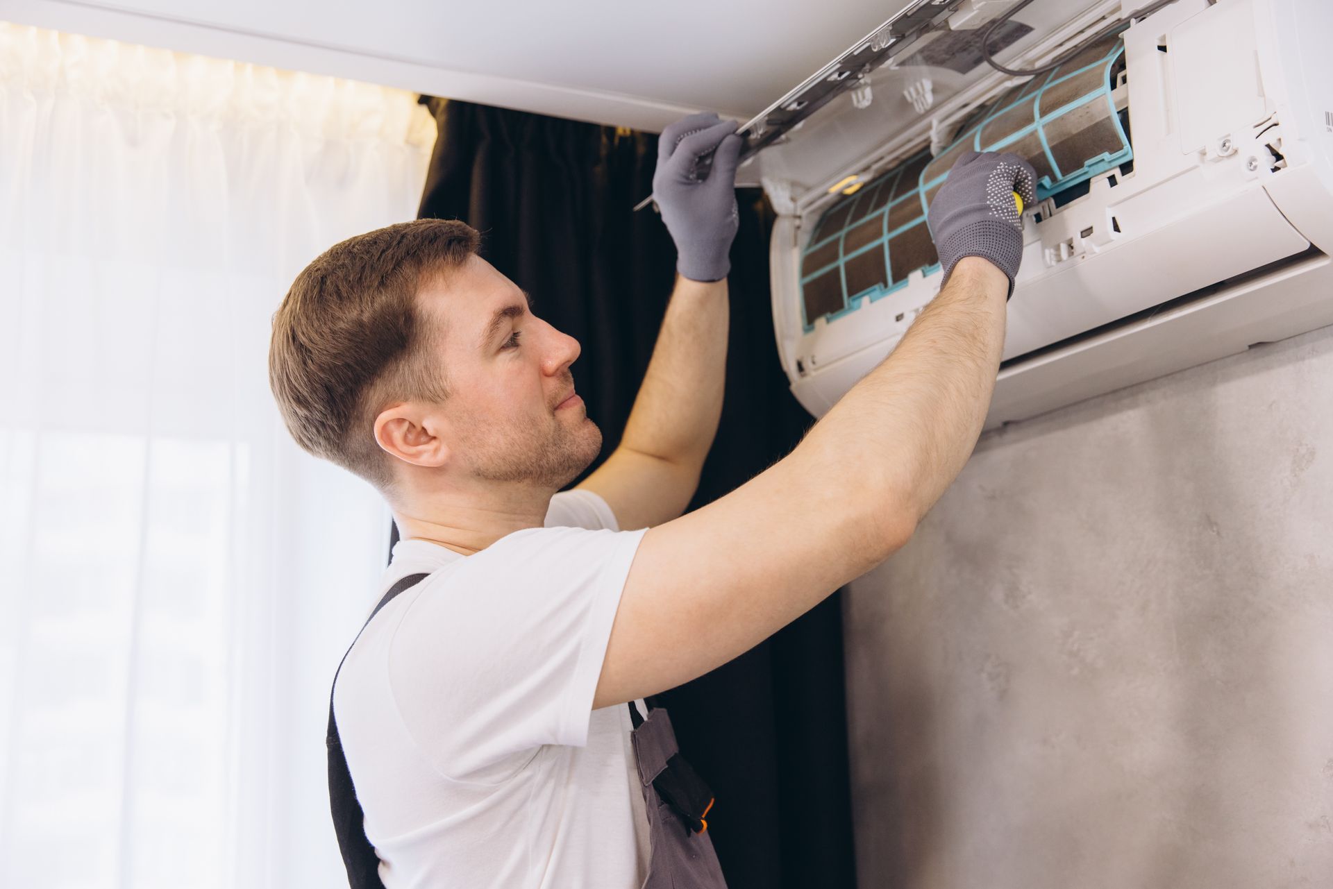 A technician wearing work gloves inspects the filter of a wall-mounted air conditioning unit indoors