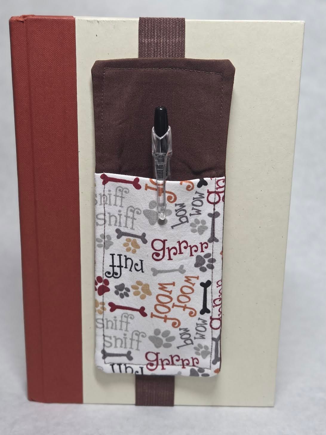 Decorative notebook cover with a burgundy spine, cream front, and patterned pocket holding a pen.