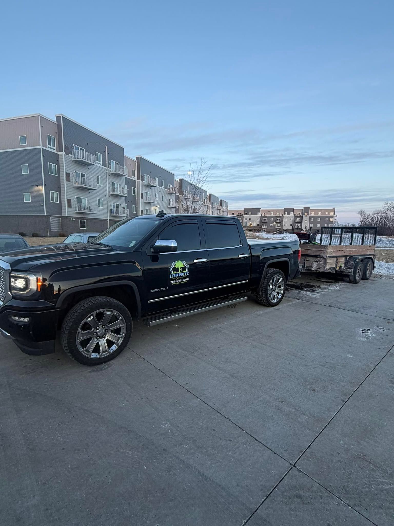 Black pickup truck towing a trailer on a roadside near apartment buildings at dusk