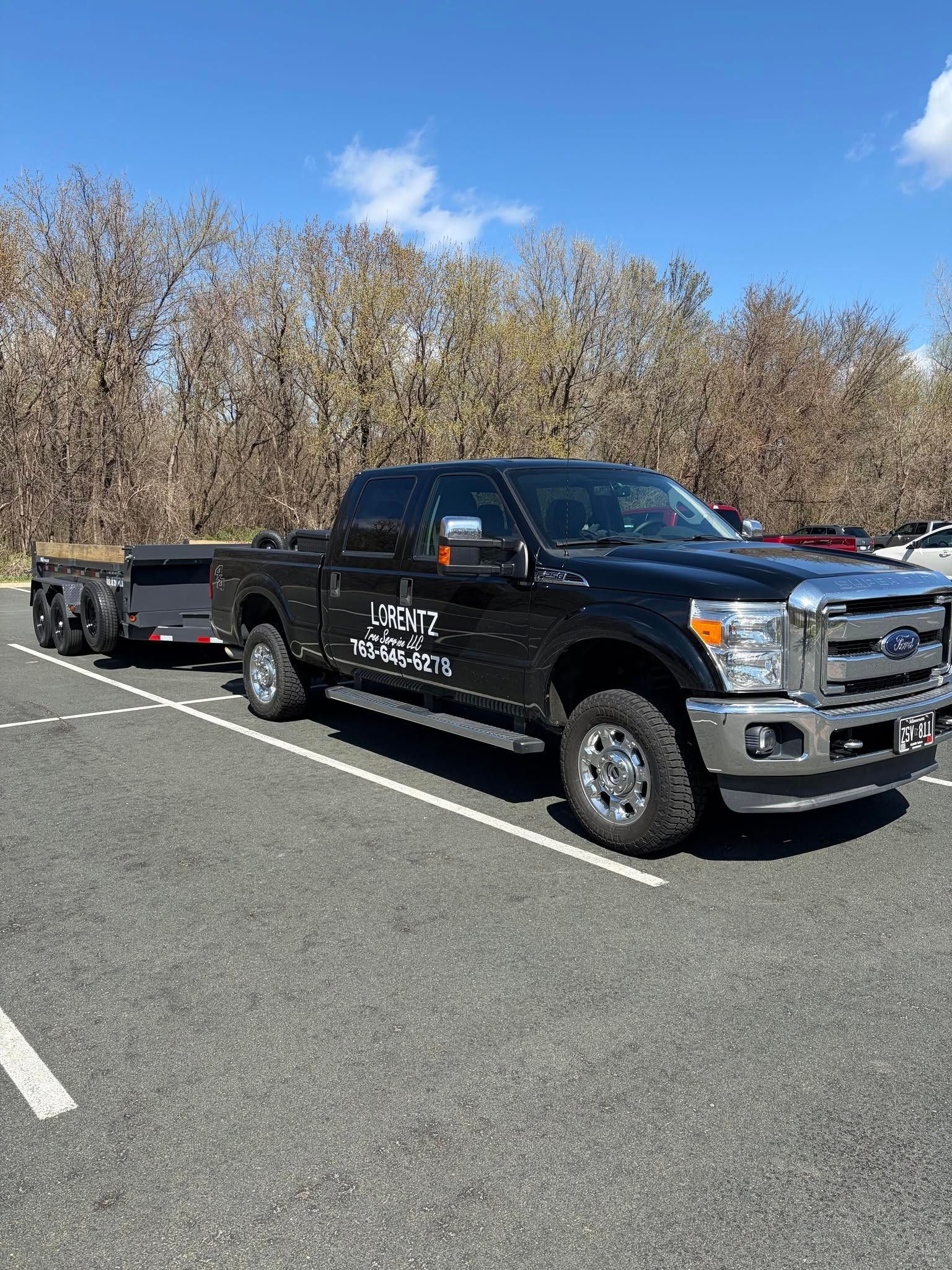 Black pickup truck with trailer parked in a lot on a sunny day, trees in the background