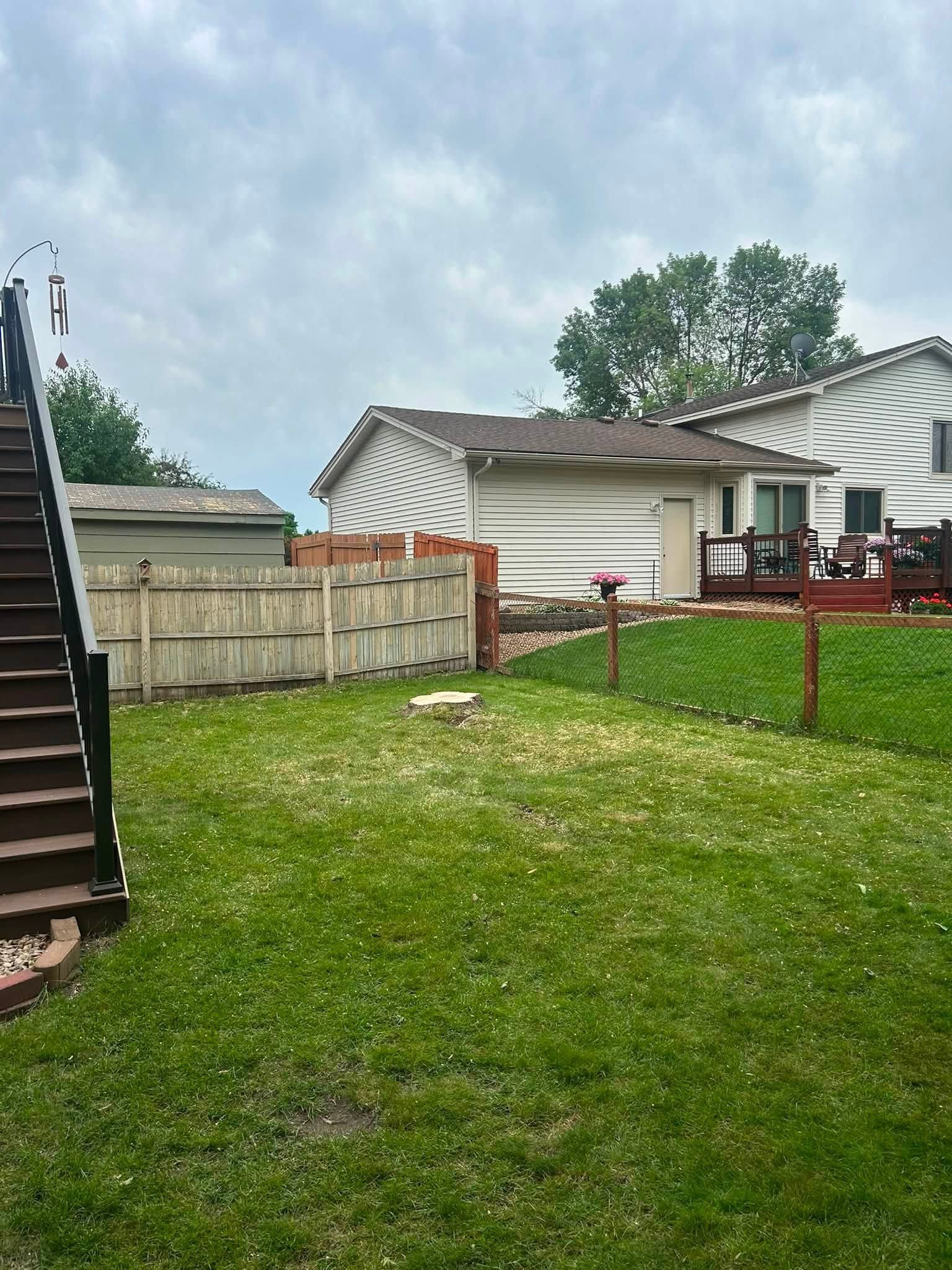 Backyard with green lawn, wooden fence, deck stairs, and patio furniture under a cloudy sky