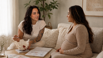 Two women talking on a couch in a bright living room, with a book and coffee table in front of them.