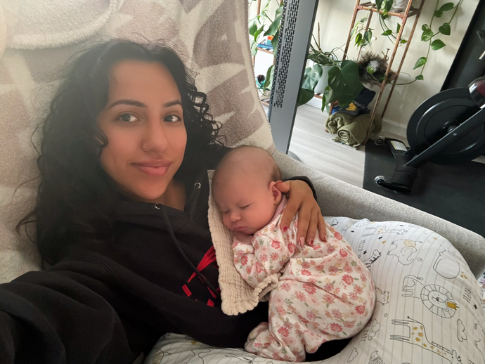 Woman holding a sleeping baby on a couch by a window, with potted plants in the background.