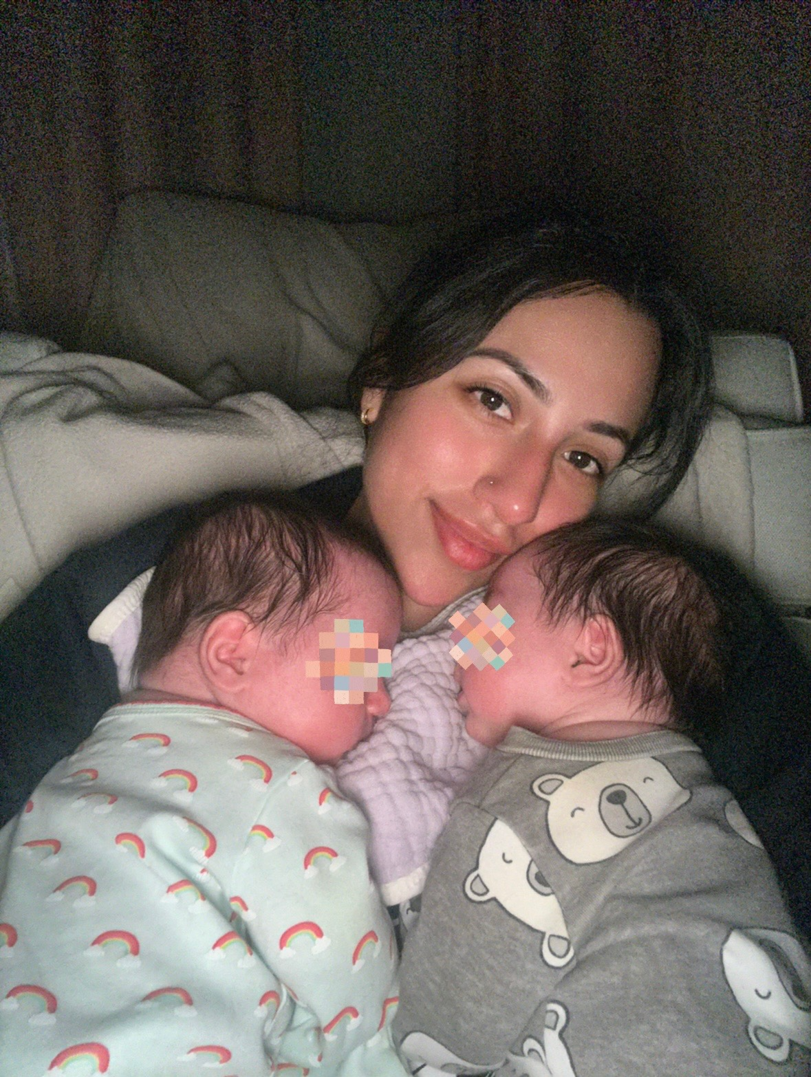 Woman lying with two sleeping babies on a bed, smiling softly in dim light.