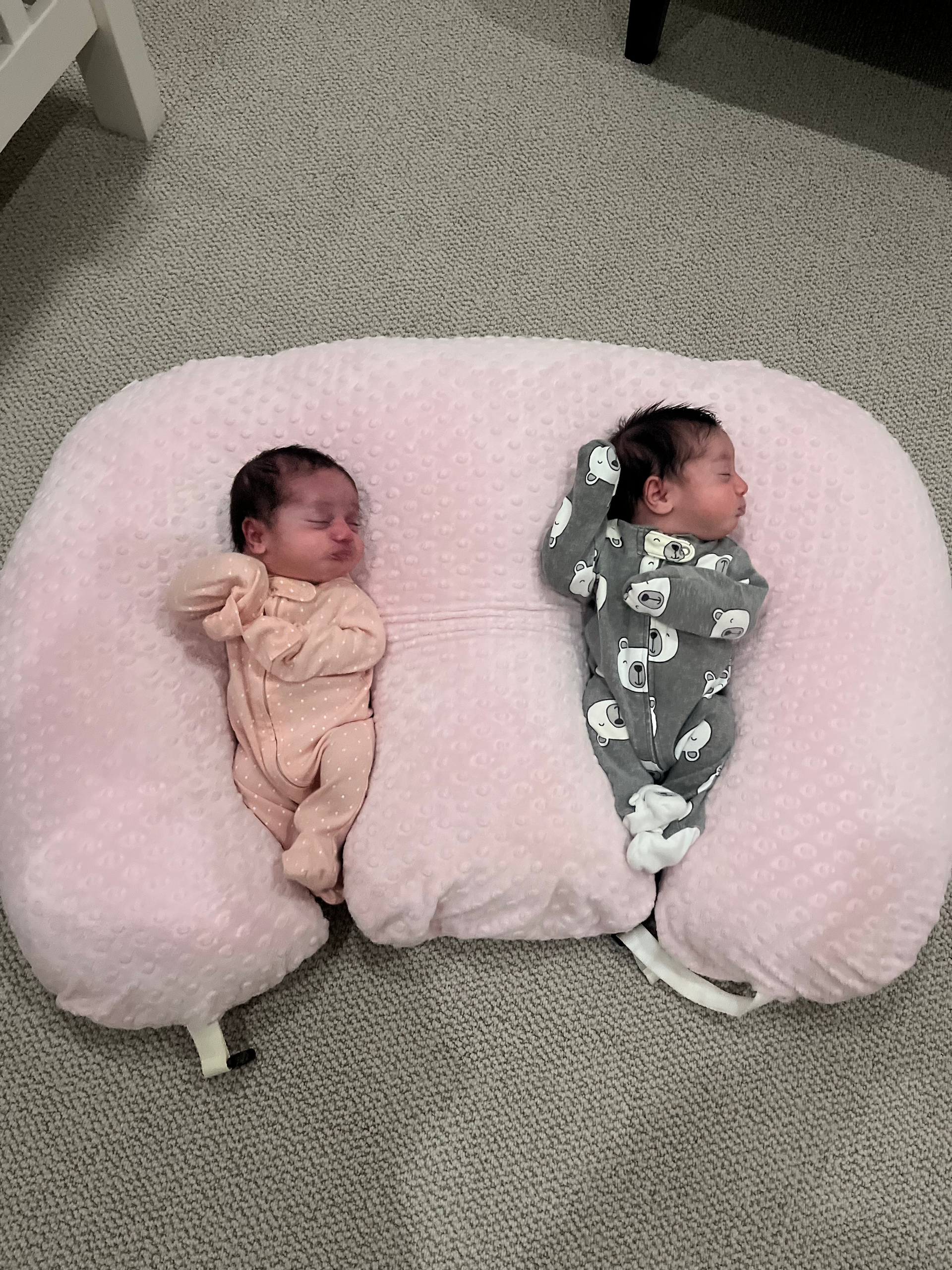 Two sleeping infants in pink and gray swaddles lying on a fluffy pink pillow on carpet.