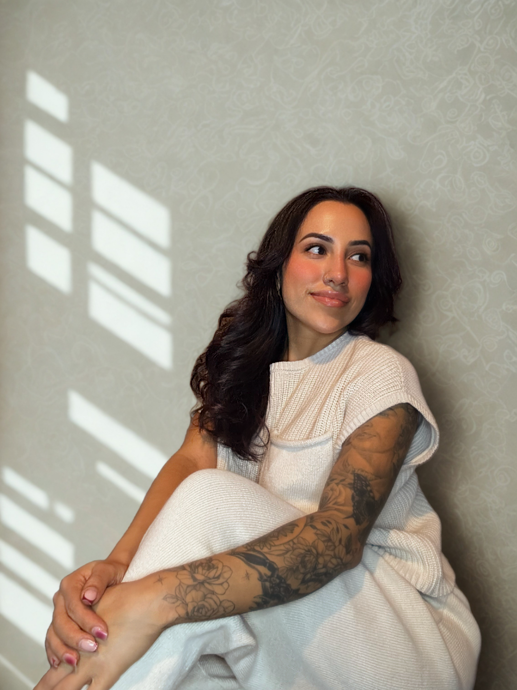 Woman in a white dress seated by a sunlit wall, with window light casting shadows behind her.