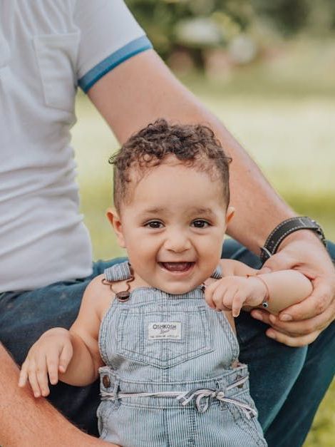 Smiling baby in denim overalls sitting outdoors, holding an adult’s hands.