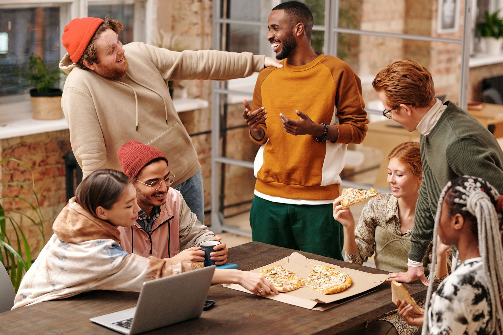 Friends gathered around a table with laptops and snacks, chatting and laughing in a casual meeting outdoors
