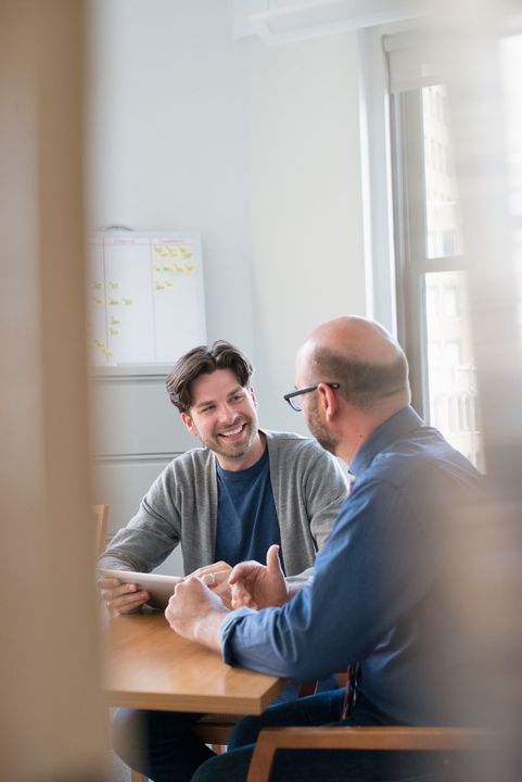 Two people talking at a wooden table in a bright office, one smiling and gesturing