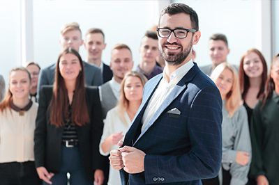 Smiling man in a blue blazer in front of a group of colleagues in an office setting.