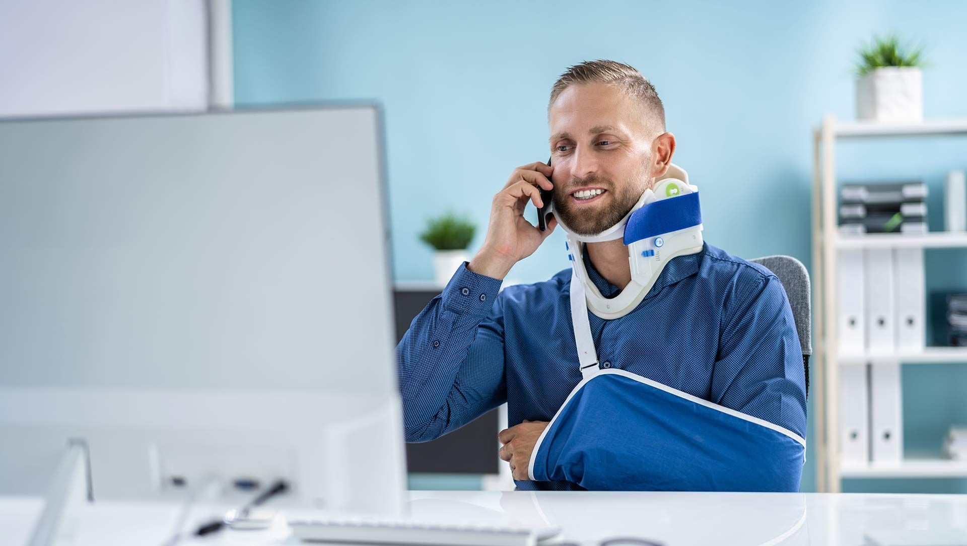 Man in a blue arm sling talking on a phone at a desk in an office