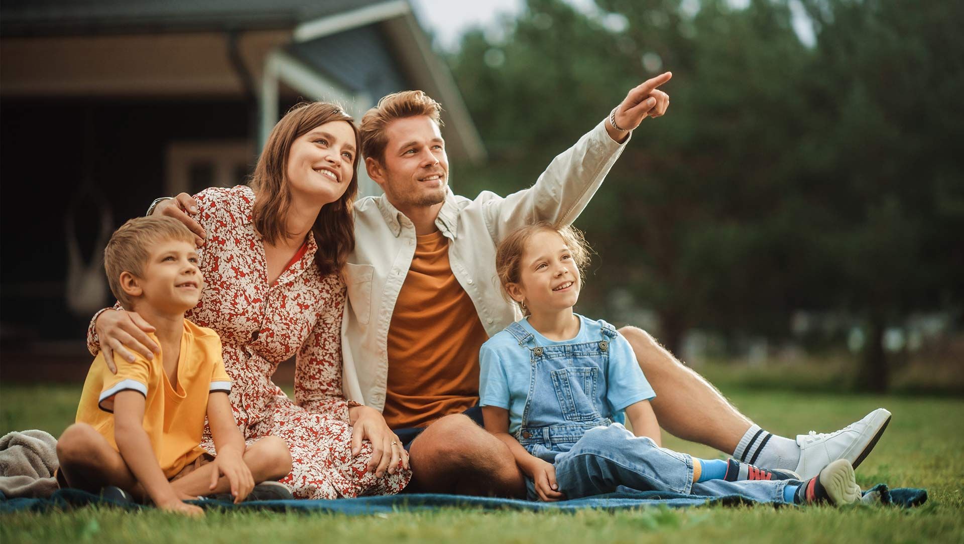 Family sitting on grass outside a house, smiling and pointing at something in the sky.