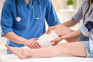 Doctor wrapping bandage on a patient's leg. Person in scrubs holds leg while another person assists. White setting.