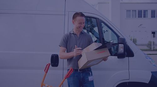 Delivery person with box next to a van. Holding box, smiling. Hand truck nearby. Outside a building.