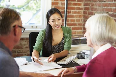 Woman in green shirt reviews documents with older couple at a desk. Brick wall background.