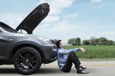 Man sits dejectedly beside a broken-down car on a roadside. Hood open, field in background.