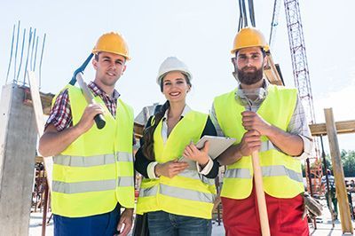 Three construction workers smiling, wearing hard hats and safety vests at a construction site.