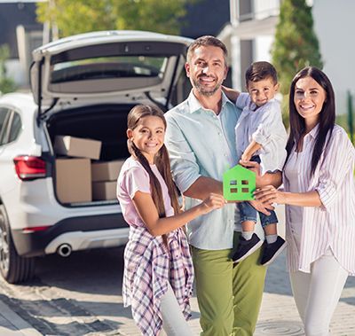 Family holding a green house cutout in front of their new home, car with boxes in the background.