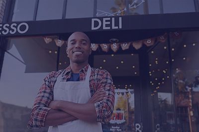 Smiling person in apron stands outside a deli, arms crossed. Espresso Deli sign is above.