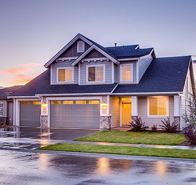 Two-story house with gray siding, dark roof, and two-car garage at dusk. Wet pavement reflects lights.