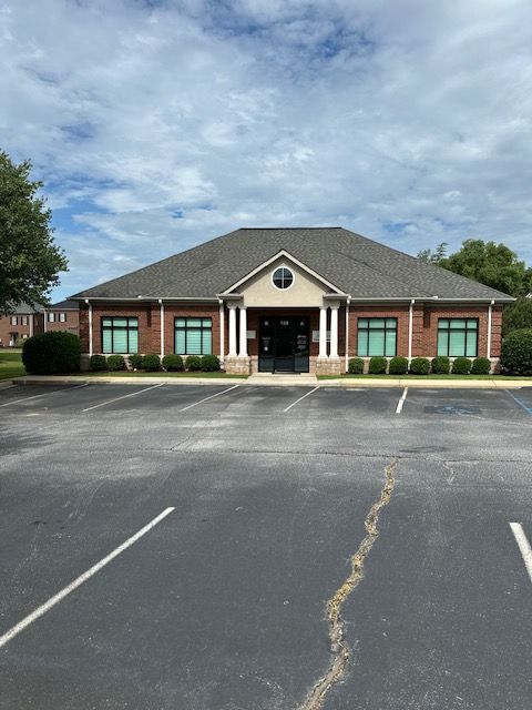 Brick building with black roof, windows, and front columns. Empty parking lot in front, cloudy sky.