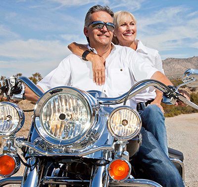 Couple on a motorcycle, smiling. Man driving, woman with arm around him. Bright sunlight.