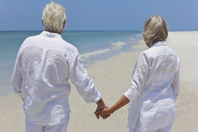 Elderly couple holding hands, walking on a beach, facing the ocean. Both wear white clothing.