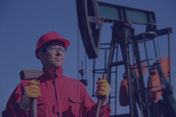 Oil worker in red overalls and helmet holding tools, standing near a working oil pump.
